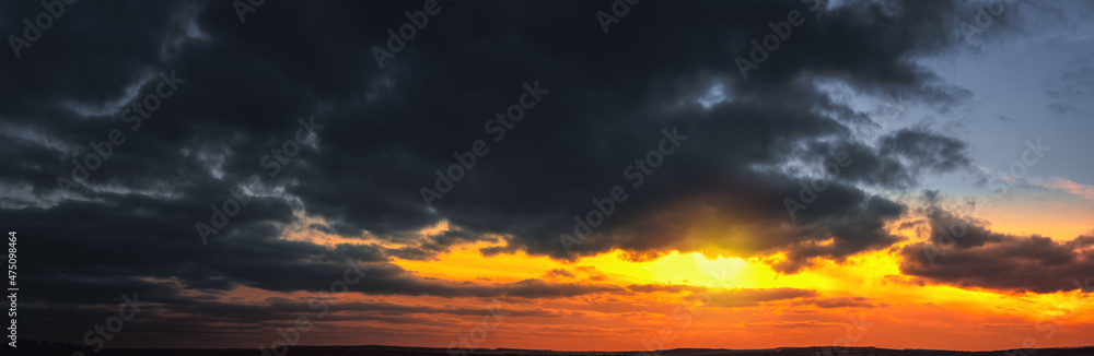 Panorama of dramatic colorful sunset with dark and bright clouds. The sun's rays are breaking through the cloudy sky. Dark clouds against a bright saturated sky.