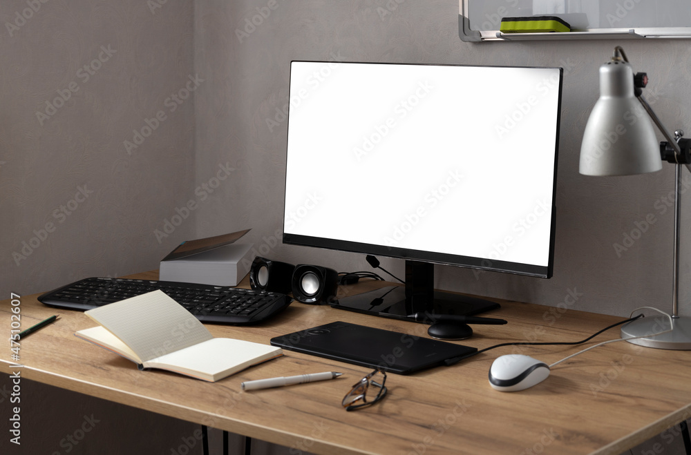 Fototapeta premium White blank screen monitor on the wooden table,tablet for drawing and designing,opened notebook and pen,mouse,lamp against white wall.Empty space
