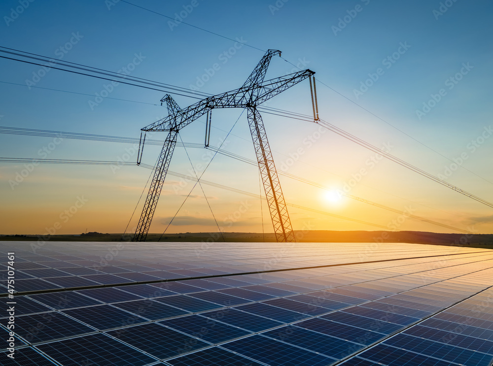 High voltage towers with electric power lines transfering energy from solar photovoltaic panels at sunset. Production of sustainable electricity concept