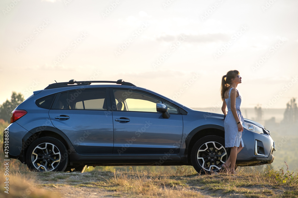 Young female driver resting near her car enjoying warm summer evening. Travel and getaway concept Young female driver resting near her car enjoying warm summer evening. Travel and getaway concept