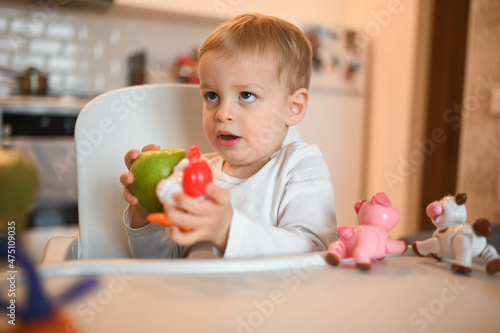 Little happy cute baby toddler boy blonde sitting on baby chair playing with apple. Baby facial expressions indoors at home kitchen interior with food. Healthy eating happy family childhood concept