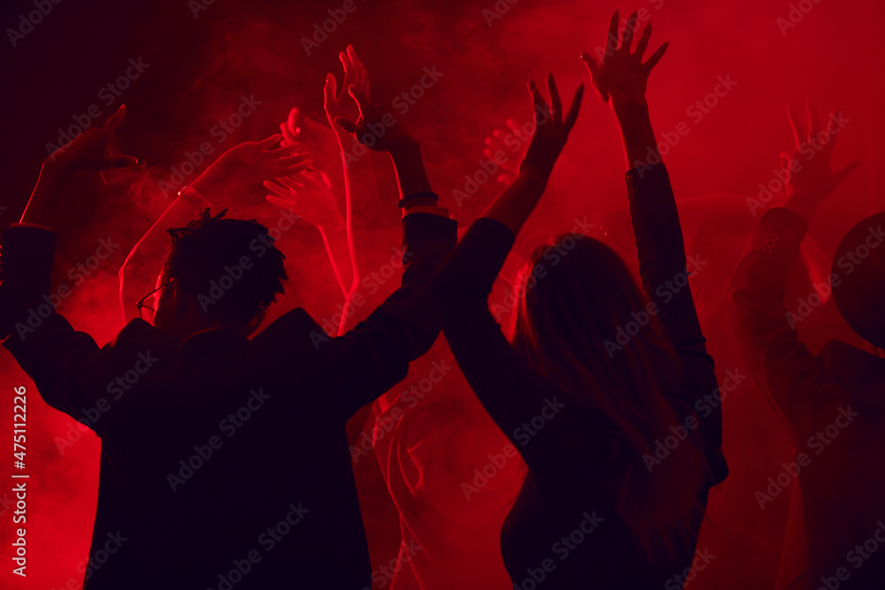 Dark silhouettes of young people dancing and raising hands while enjoying party in smoky nightclub lit by red light, copy space