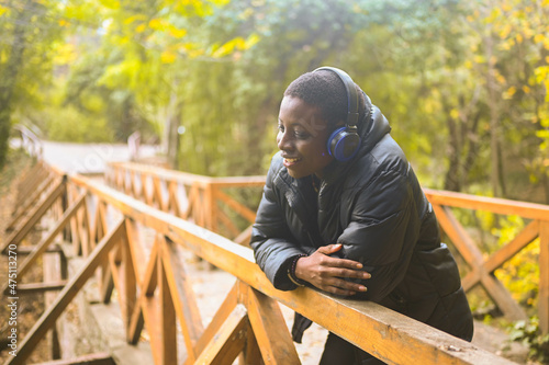 Attractive happy smiling young natural beauty short haired African Black woman with blue headphones in black down jacket leans on a wooden railing listening music in nature summer park