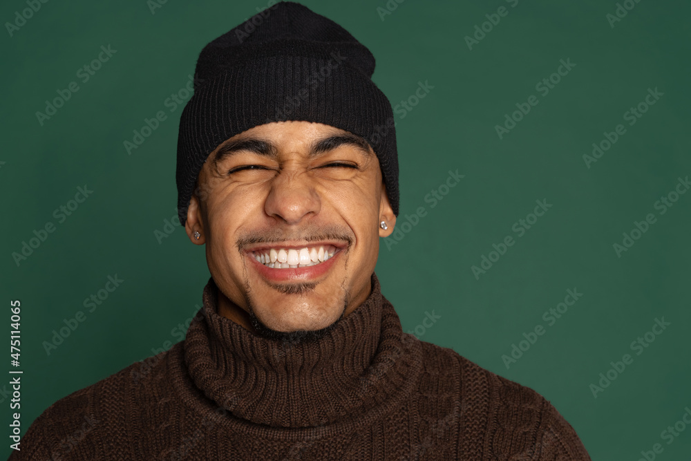 Close-up portrait of cheerful African young man in warm clothes and hat isolated on green color studio background. Concept of human emotions Close-up portrait of cheerful African young man in warm clothes and hat isolated on green color studio background. Concept of human emotions