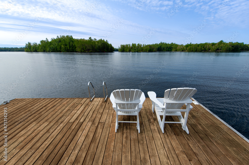 Muskoka chairs sitting on a cottage wooden dock facing the blue water of a lake in Ontario Canada during the summer.