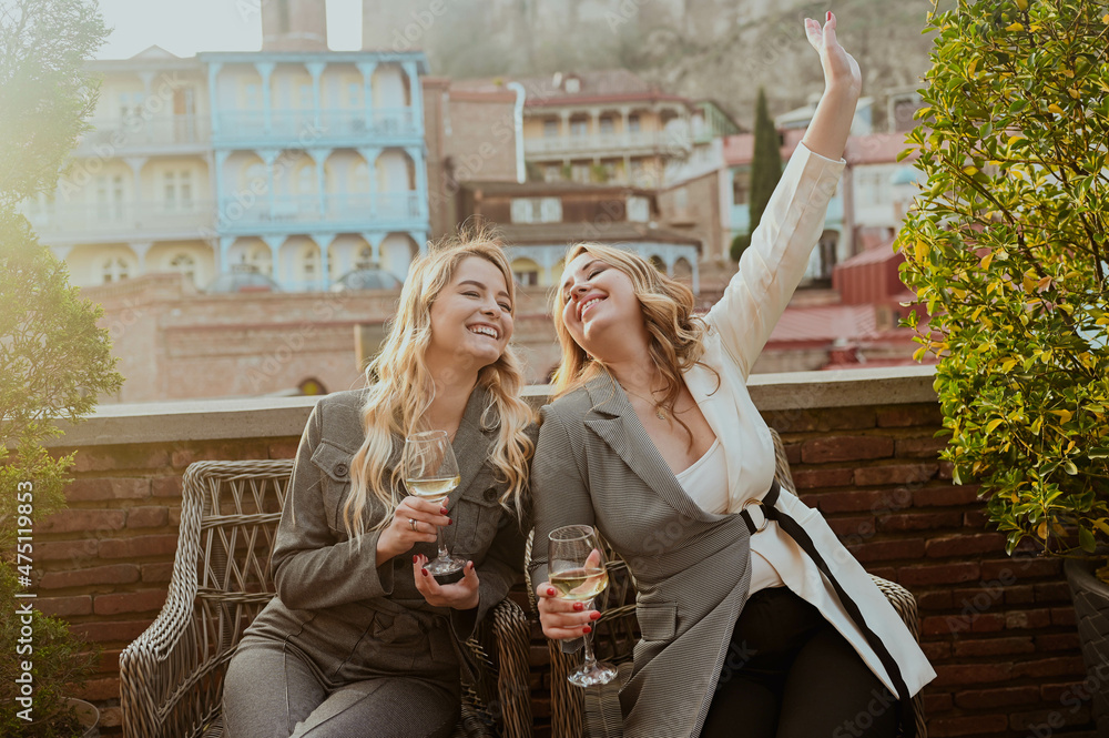 Close-up portrait of two female friends in strict suits laughing drinking wine on the terrace outside at summer street cafe on background buildings of Old Tbilisi city, Georgia. High quality photo