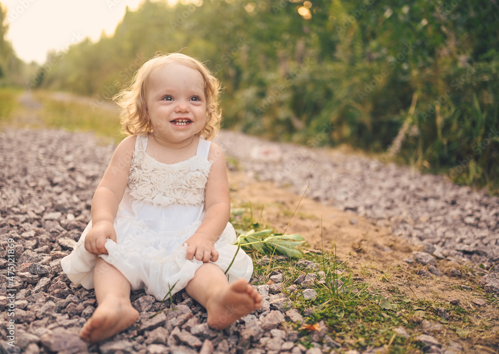 Little funny cute blonde girl child toddler with curls in white dress and with mud on her face sits on the ground on the street outside at summer. Baby laughing. Happy healthy childhood concept Little funny cute blonde girl child toddler with curls in white dress and with mud on her face sits on the ground on the street outside at summer. Baby laughing. Happy healthy childhood concept