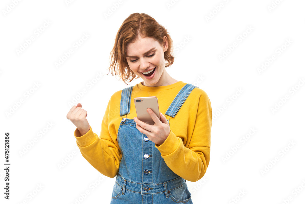 lucky smiling redhead girl holding smartphone in yellow clothes isolated on white studio background received joyful message, celebrating victory