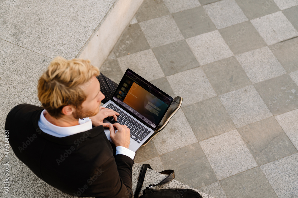 Obraz premium Blonde man working with laptop while sitting on stone bench