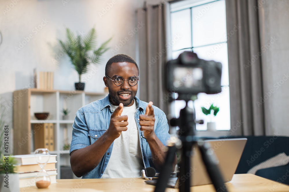 Positive young guy recording video blog on camera while staying at home. African american man in eyewear and casual outfit creating new content for his social networks. - obrazy, fototapety, plakaty 