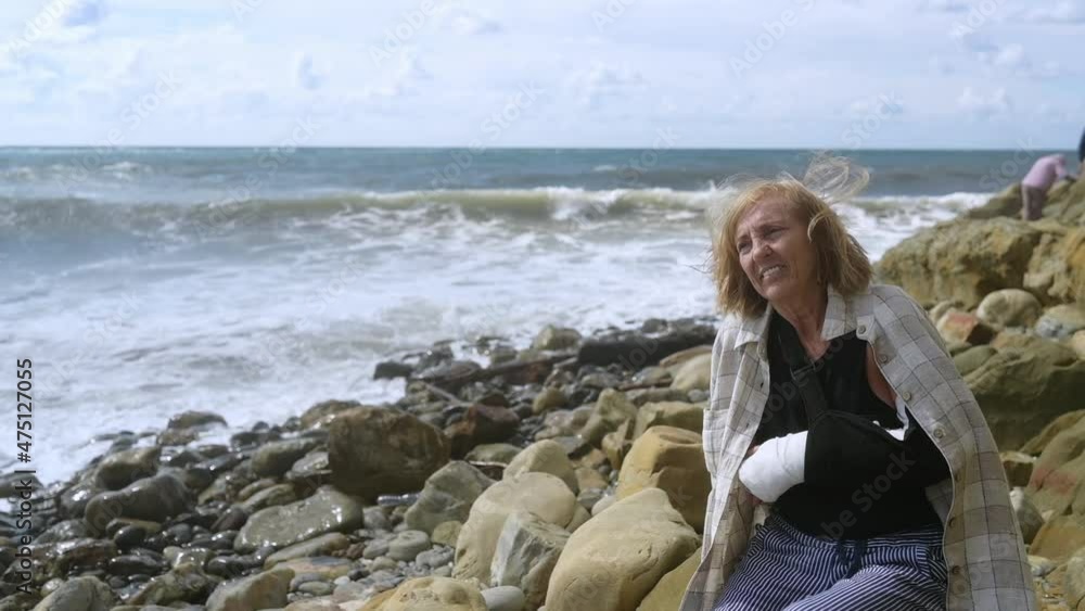 Senior elderly woman relaxing sitting on coast of ocean or sea windy cloudy day suffering from joint pain. Injury broken arm hand. Old lady wrist in gypsum sling bandage feeling hurt outside in summer Senior elderly woman relaxing sitting on coast of ocean or sea windy cloudy day suffering from joint pain. Injury broken arm hand. Old lady wrist in gypsum sling bandage feeling hurt outside in summer