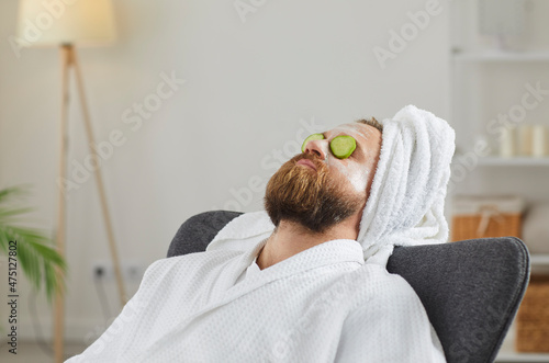 Adult man in bathrobe and towel turban enjoying spa day at home and relaxing in comfortable chair or armchair with facial beauty mask on face and fresh cucumber slices on eyes. Skin care concept photo