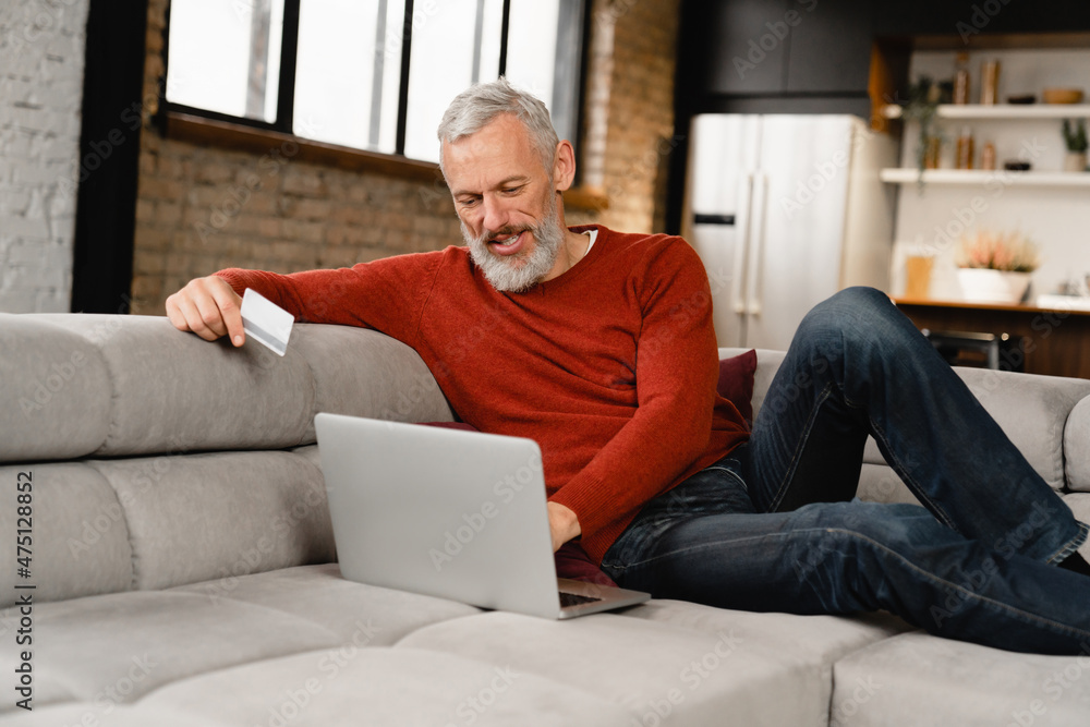 Smiling caucasian mature middle-aged man doing shopping online, buying purchasing presents clothes remotely, ordering food delivery with credit card e-commerce banking on laptop at home Smiling caucasian mature middle-aged man doing shopping online, buying purchasing presents clothes remotely, ordering food delivery with credit card e-commerce banking on laptop at home
