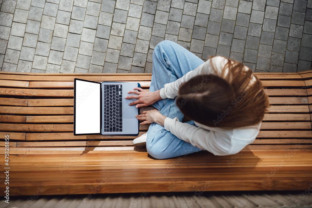 Top view, close-up of smartphone with blank screen in hands of young woman sitting at wooden bench and touching screen. Laptop with blank screen. Mock up space for ad.