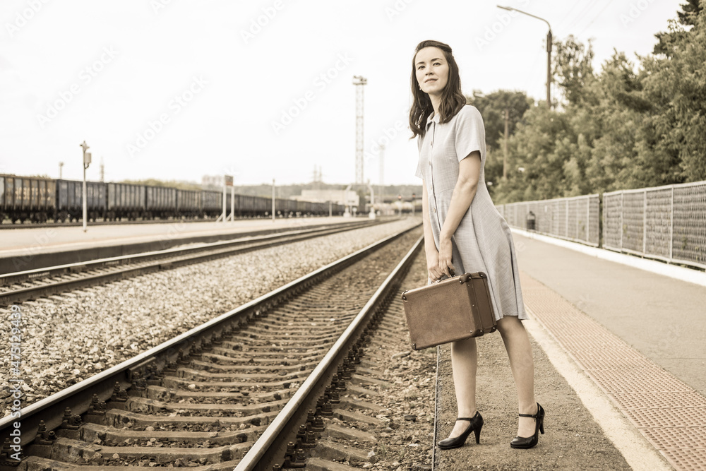 A young woman in a retro dress with a vintage suitcase in her hands is standing on the railway platform and waiting for the arrival of the train. A woman is waiting for a train.