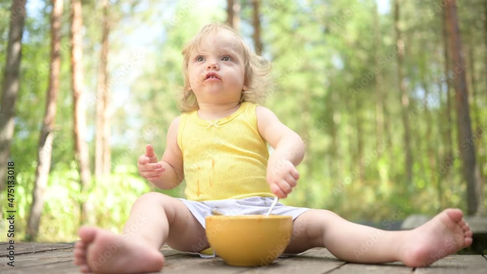 Little funny cute blonde girl child toddler with dirty clothes and face eating baby food fruit or vegetable puree with spoon from yellow plate outside at summer. Healthy happy childhood concept Little funny cute blonde girl child toddler with dirty clothes and face eating baby food fruit or vegetable puree with spoon from yellow plate outside at summer. Healthy happy childhood concept