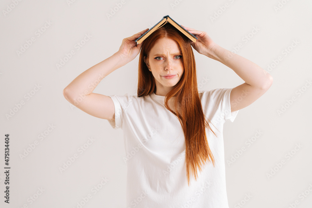 Studio portrait of angry young student woman covering head with book looking at camera, standing on white isolated background. Confused redhead lady posing with educational materials. Studio portrait of angry young student woman covering head with book looking at camera, standing on white isolated background. Confused redhead lady posing with educational materials.