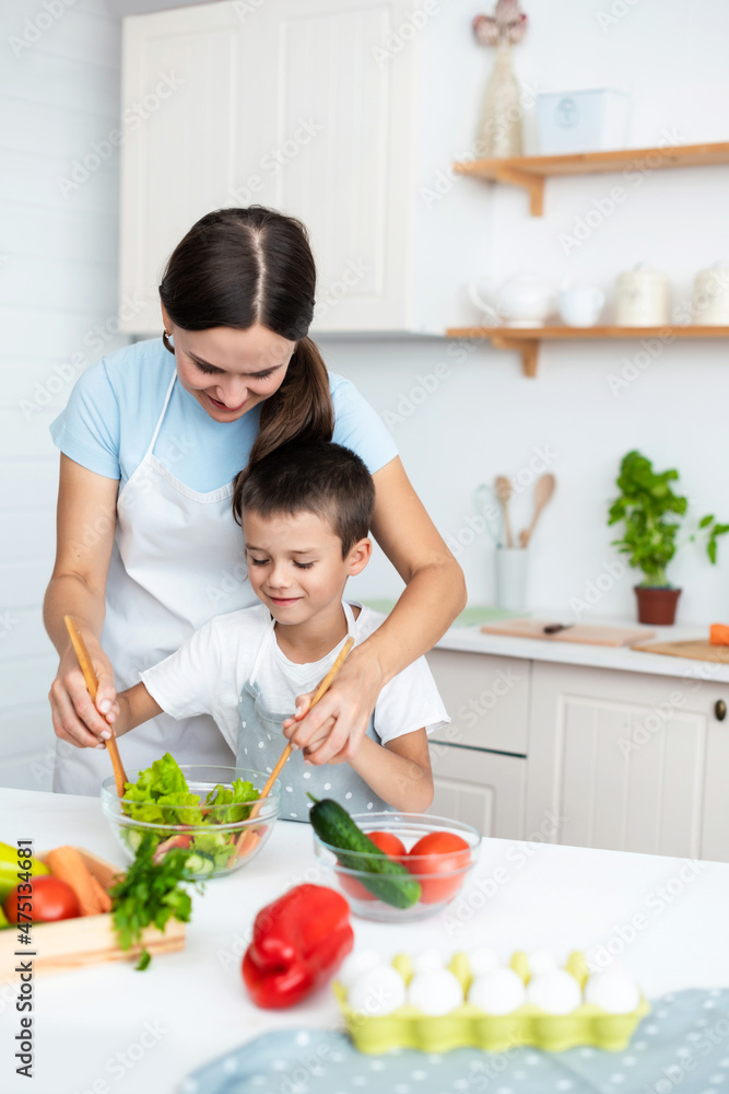 Bonding Concept. Cheerful mother helping her son to cook, teaching little boy how to prepare healthy vegetable salad, mixing ingredients in bowl, cooking together at home.