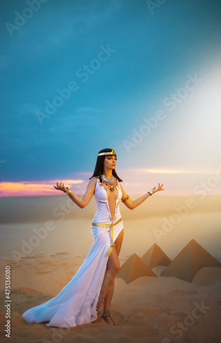 Egypt Woman pagan prays hands raised to heaven blue sky. Sexy girl Egyptian goddess Queen Cleopatra. yellow sand Sahara desert pyramids. Art ancient pharaoh costume white dress gold accessories crown. photo