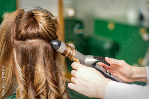 Creating curls with curling irons. Hairdresser makes a hairstyle for a young woman with long red hair in a beauty salon