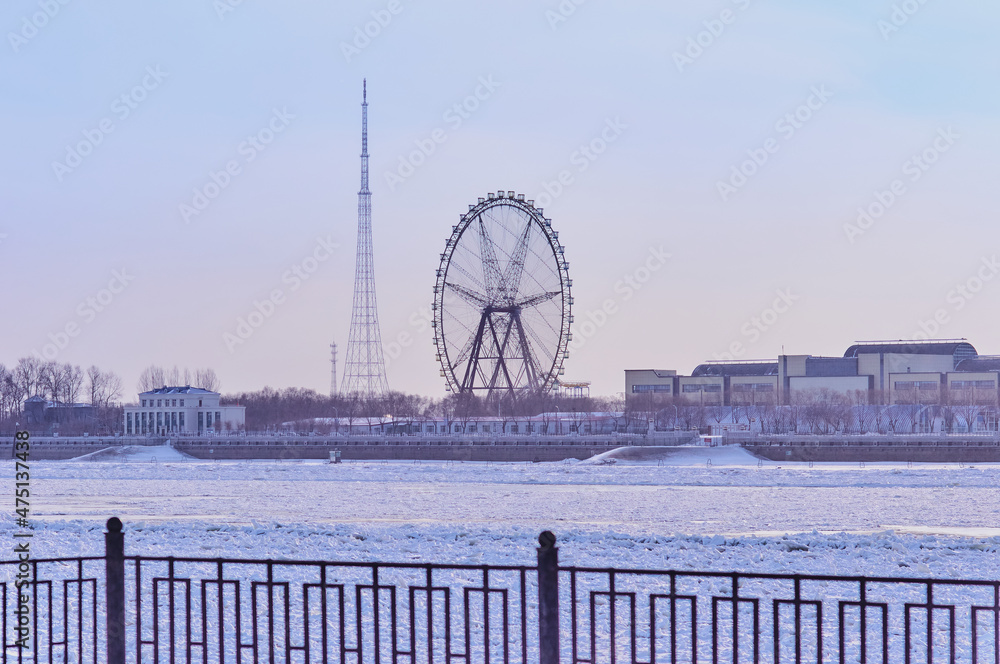 View of the Chinese Amusement Park with a Ferris wheel from the city of Blagoveshchensk, Russia through the embankment fence. Winter morning. The Amur River is ice-bound.