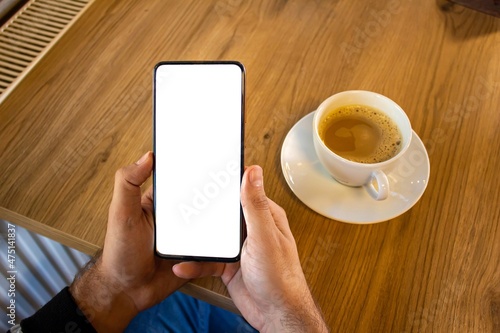 Hand holding mobile phone with blank white screen while drinking coffee.