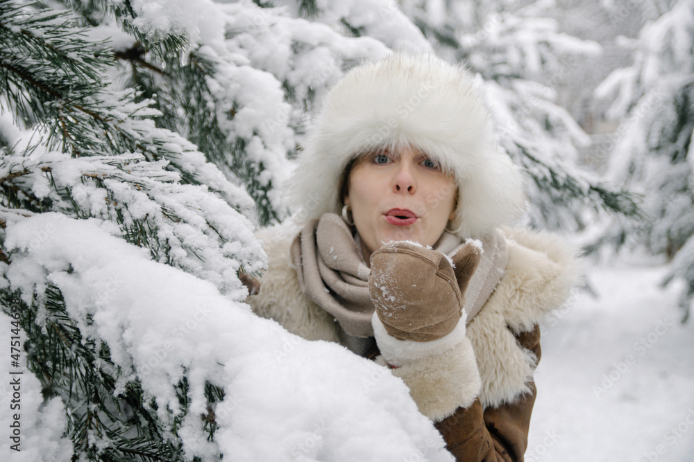 horizontal winter portrait of a smiling young European girl in a sheepskin coat, mittens, a white hat with earflaps and boots, who stands in snow-covered pines and blows snow from mittens horizontal winter portrait of a smiling young European girl in a sheepskin coat, mittens, a white hat with earflaps and boots, who stands in snow-covered pines and blows snow from mittens
