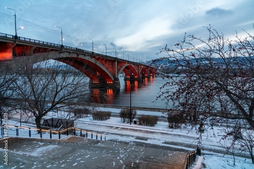 View of the Communal Bridge (1961) with night illumination across the Yenisei River from the embankment of the right bank in the city of Krasnoyarsk. Russia.
