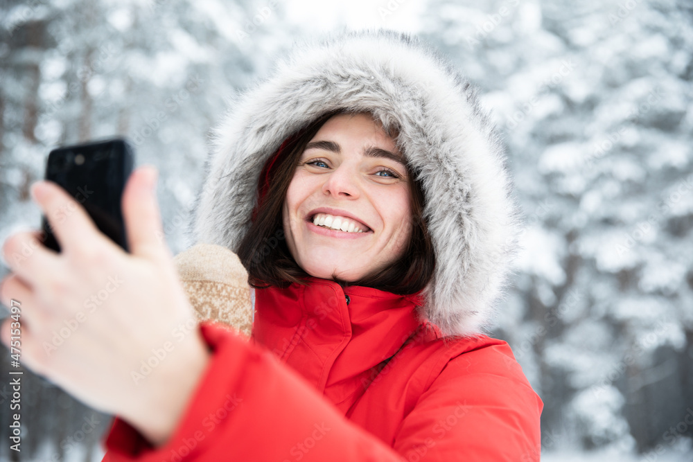 Portrait of beautiful young laughing brunette with gray eyes in red jacket with hood made of faux fur on winter day against background of snowy trees Woman takes selfie on her mobile phone and talks Portrait of beautiful young laughing brunette with gray eyes in red jacket with hood made of faux fur on winter day against background of snowy trees Woman takes selfie on her mobile phone and talks