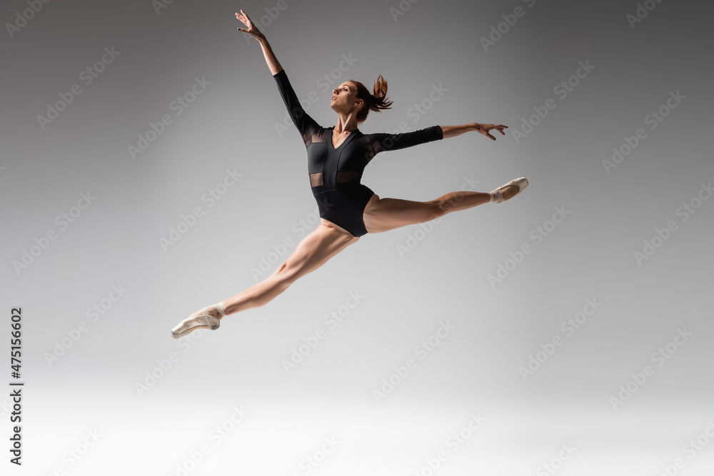 full length of young ballerina in pointe shoes and black bodysuit jumping on dark gray