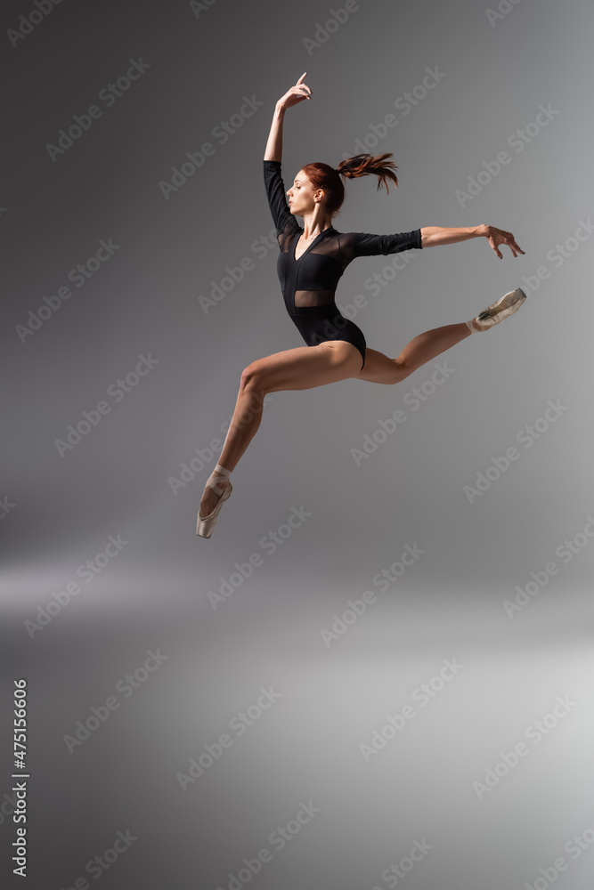 pretty ballerina in pointe shoes and black bodysuit jumping on dark grey