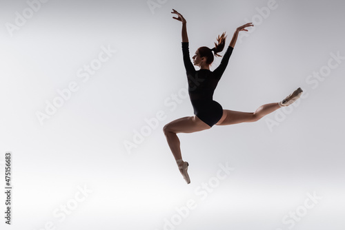 Fototapeta Naklejka Na Ścianę i Meble -  full length of young ballerina in black bodysuit levitating on grey