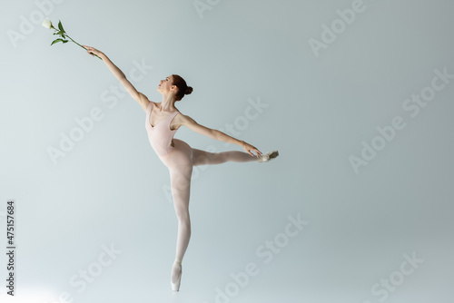 Fotografia full length of graceful ballerina in bodysuit holding rose and dancing on grey
