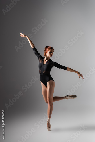 graceful ballerina in pointe shoes and black bodysuit with outstretched hands on dark grey