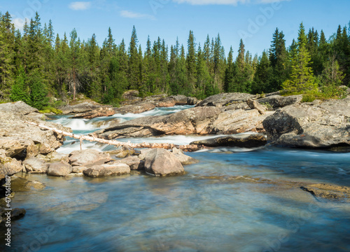 Beautiful landscape with long exposure water stream and cascade of river Kamajokk, boulders and spruce tree forest in Kvikkjokk village in Swedish Lapland. Summer sunny day, blue sky, white clouds