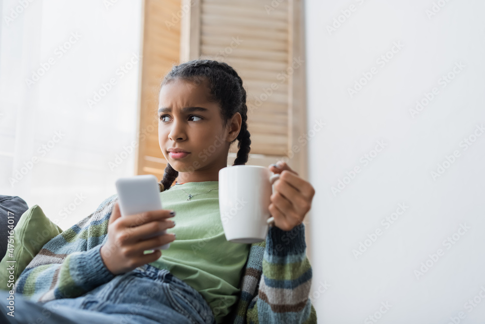 upset african american teenager holding cup of tea and blurred mobile phone at home upset african american teenager holding cup of tea and blurred mobile phone at home
