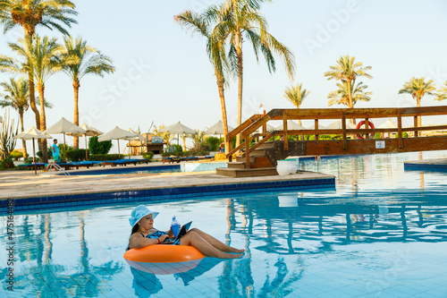 A brunette woman in a sun hat in a swimming pool in a swimming circle with a cocktail and a laptop communicates by video call on a background of palm trees.