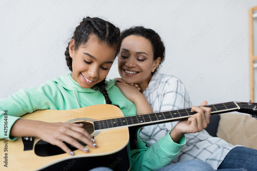 happy african american woman smiling near teenage daughter playing guitar at home happy african american woman smiling near teenage daughter playing guitar at home