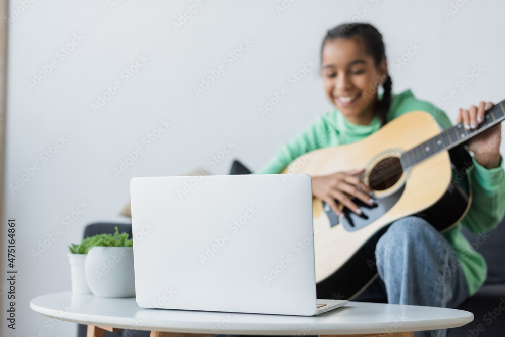 blurred african american teenage girl smiling while learning to play acoustic guitar near laptop blurred african american teenage girl smiling while learning to play acoustic guitar near laptop