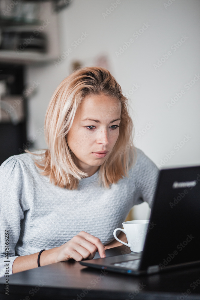 Female freelancer in her casual home clothing working remotly from her dining table in the morning. Home kitchen in the background.