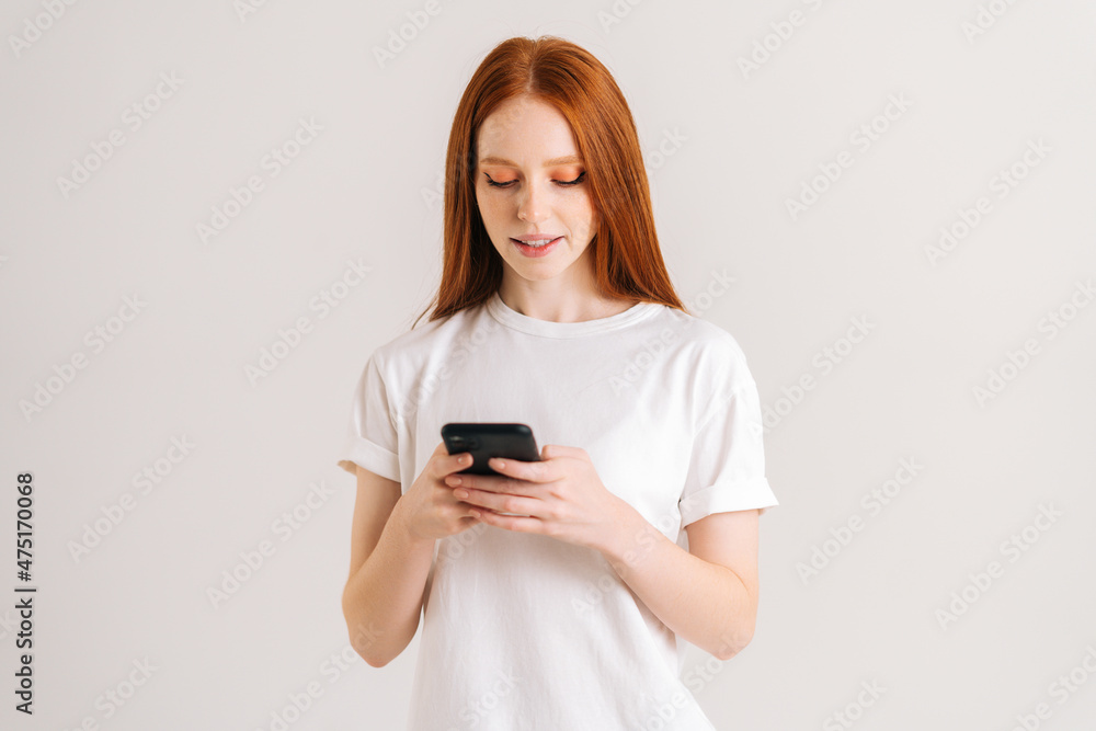 Studio portrait of attarctive young woman typing online message using mobile phone on white isolated background. Smiling redhead lady thinking over answer on social media and texting on smartphone Studio portrait of attarctive young woman typing online message using mobile phone on white isolated background. Smiling redhead lady thinking over answer on social media and texting on smartphone
