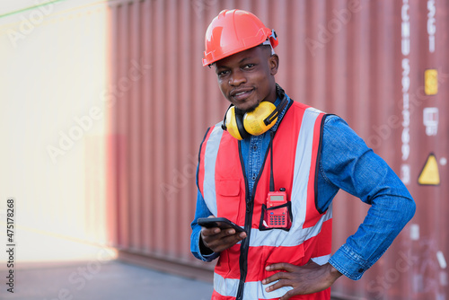 African black male worker standing smiling happily holding smartphone and wearing safety work equipment clothes Work at a warehouse, ship, cargo, import, export industry.