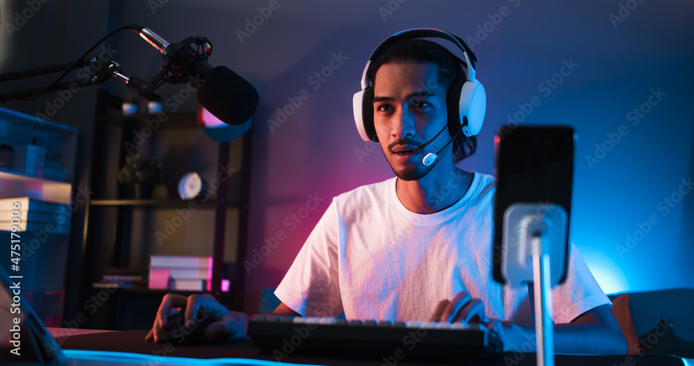 Young confident Asian man playing online computer video game, colorful lighting broadcast streaming live at home. Gamer lifestyle, E-Sport online gaming technology concept - obrazy, fototapety, plakaty 