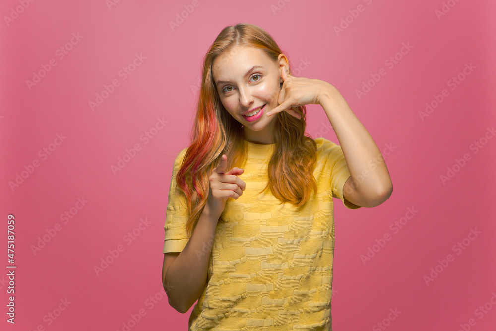 Happy young girl show call me gesture pointing finger at camera, offer communication connection on pink background Happy young girl show call me gesture pointing finger at camera, offer communication connection on pink background