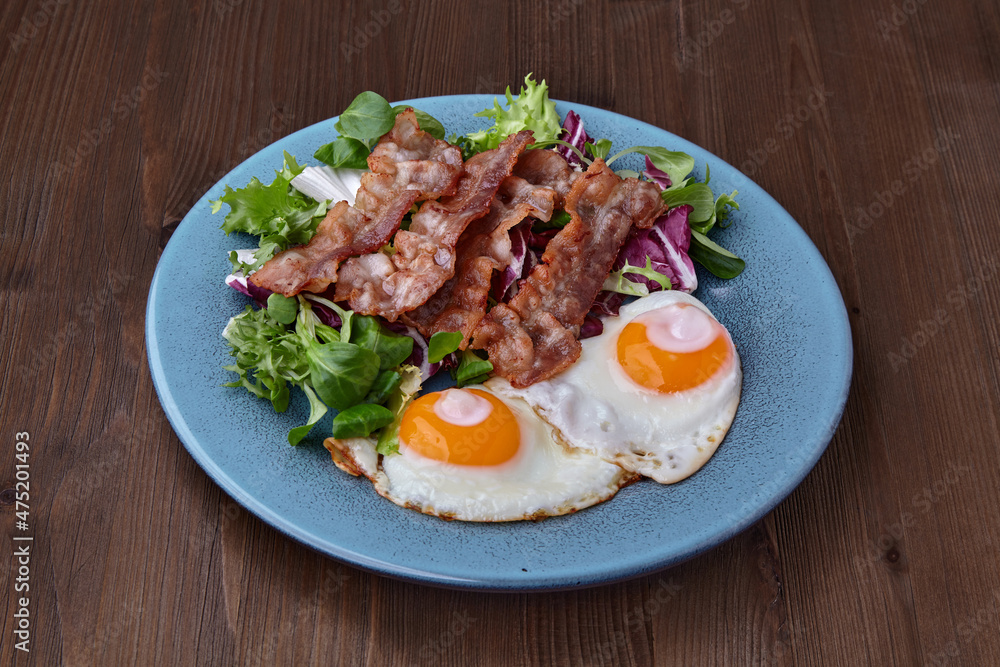 Fried eggs on a blue plate with bacon and salad on a brown wooden table