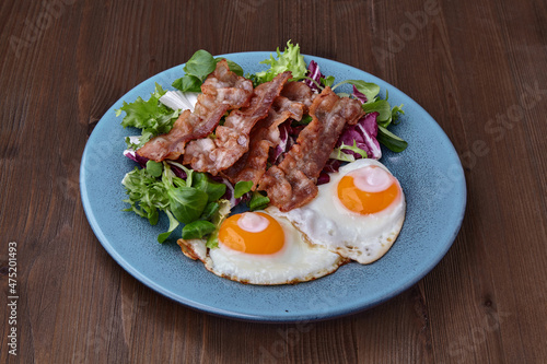 Fried eggs on a blue plate with bacon and salad on a brown wooden table