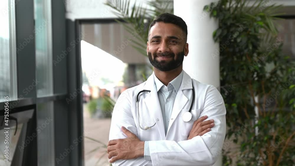 Handsome smart, proud Indian professional therapist in medical uniform and stethoscope, standing in hospital on blurred background looking at camera and smiling friendly. Healthcare, medicine concept Handsome smart, proud Indian professional therapist in medical uniform and stethoscope, standing in hospital on blurred background looking at camera and smiling friendly. Healthcare, medicine concept
