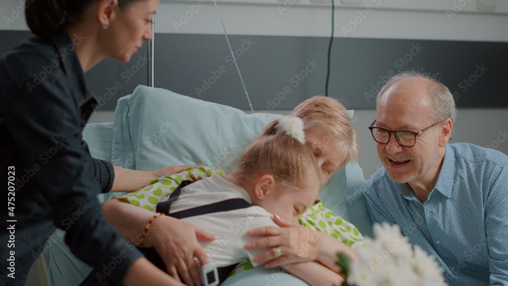 Obraz premium Close up of ill pensioner receiving visit from daughter and niece in hospital ward. Child and mother bringing flowers and visiting senior woman with disease. Visitors giving comfort to patient