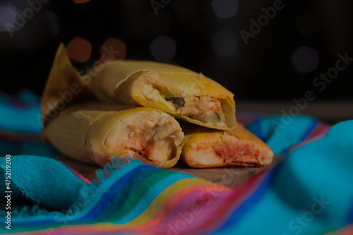 Photo of some delicious tamales, made of red mole, with a bokeh of Christmas lights in the background and a blue fabric with Mexican pink and yellow stripes.
