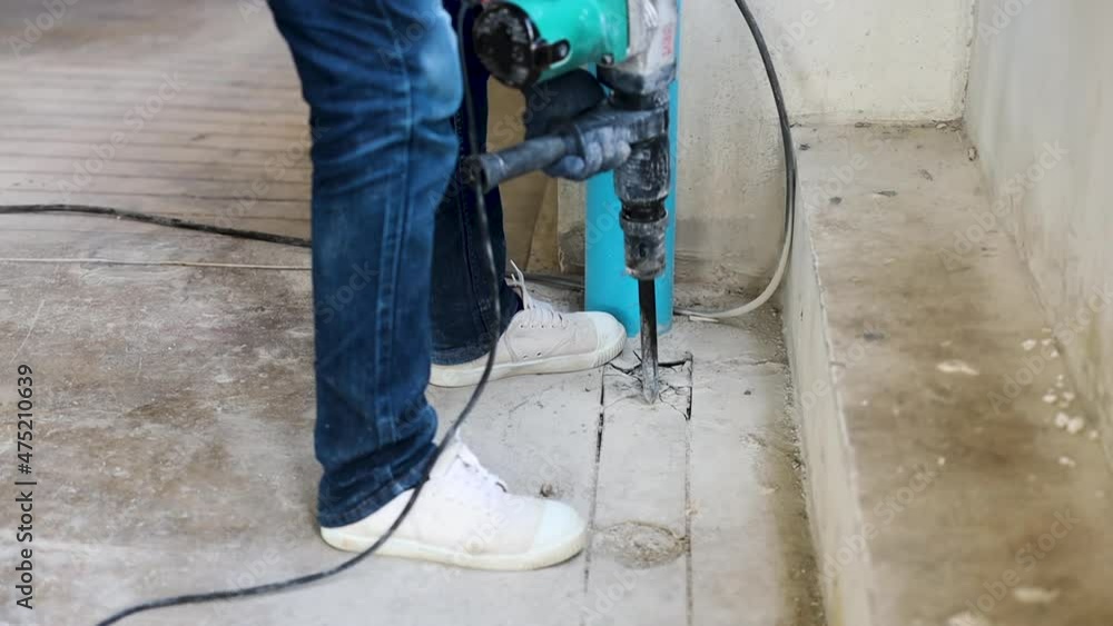 A construction worker is using a large drill to drill holes in the cement floor to make a drainage channel in a construction site. A construction worker is using a large drill to drill holes in the cement floor to make a drainage channel in a construction site.